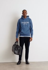 Hoodie in cotone blu con logo bianco, pantaloni neri e borsa da viaggio in pelle nera. Texture liscia, design moderno e dettagli pratici.