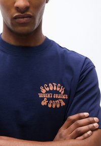Mand iført mørkeblå T-shirt med orange tekst "Scotch Desert Solstice & Soda" på venstre bryst, armene krydset over torsoen.