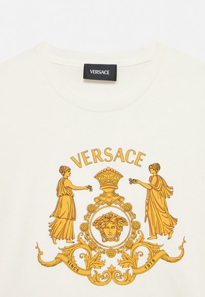 T-shirt crème avec un emblème doré de style classique montrant deux femmes drapées offrant des fleurs autour d'une tête de Méduse couronnée, texte "VERSACE" et "SINCE 1978".