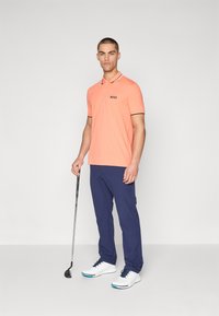 Polo arancione con logo BOSS, dettagli neri. Pantaloni blu navy e scarpe da golf bianche con dettagli blu. Mazza da golf in mano, sfondo neutro.