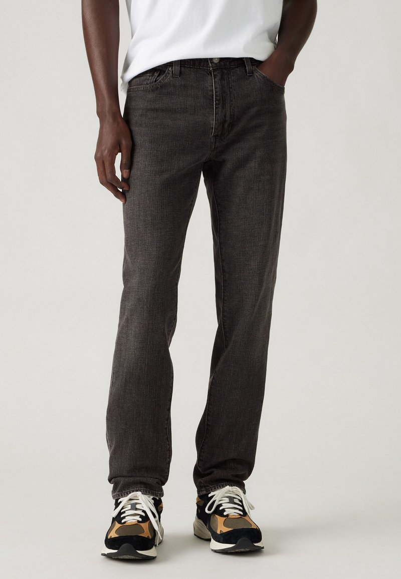 Jeans gris oscuro de corte recto, con una textura sutil y un diseño clásico de cinco bolsillos, combinados con zapatillas negras y marrones.