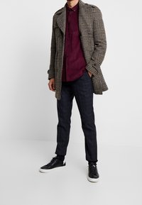 Karierter Mantel in Braun und Beige, dunkelrotes Hemd mit Knopfleiste, dunkle karierte Hose, schwarze Sneaker mit weißen Sohlen. Modell in stehender Pose.