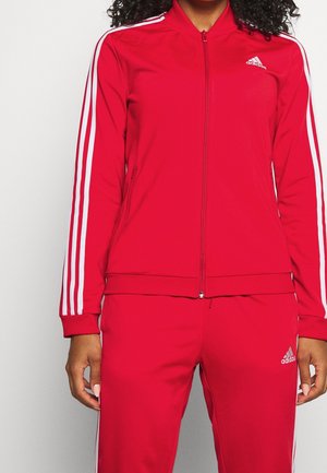 Persoon draagt een rood Adidas trainingspak met witte strepen op de mouwen en broek, staand tegen een effen lichte achtergrond.