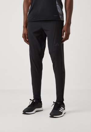 Mężczyzna w czarnych spodniach sportowych z logo Adidas na udzie oraz czarnych butach do biegania z białymi podeszwami i paskami, stojący na jednolitym tle.