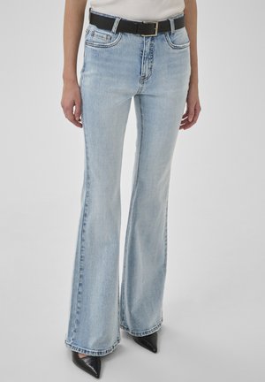 Donna con jeans svasati azzurri, cintura nera, camicia bianca infilata e scarpe nere con punta, in piedi contro uno sfondo semplice.