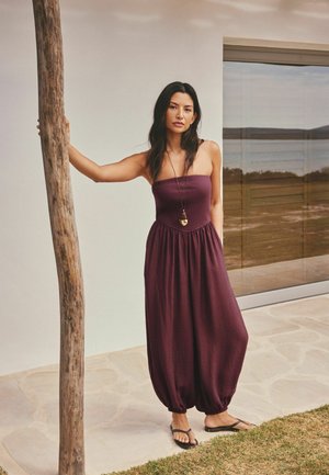 Vrouw in een strapless bordeaux jumpsuit en zwarte sandalen leunt op een houten paal voor een modern gebouw met grote glasramen.