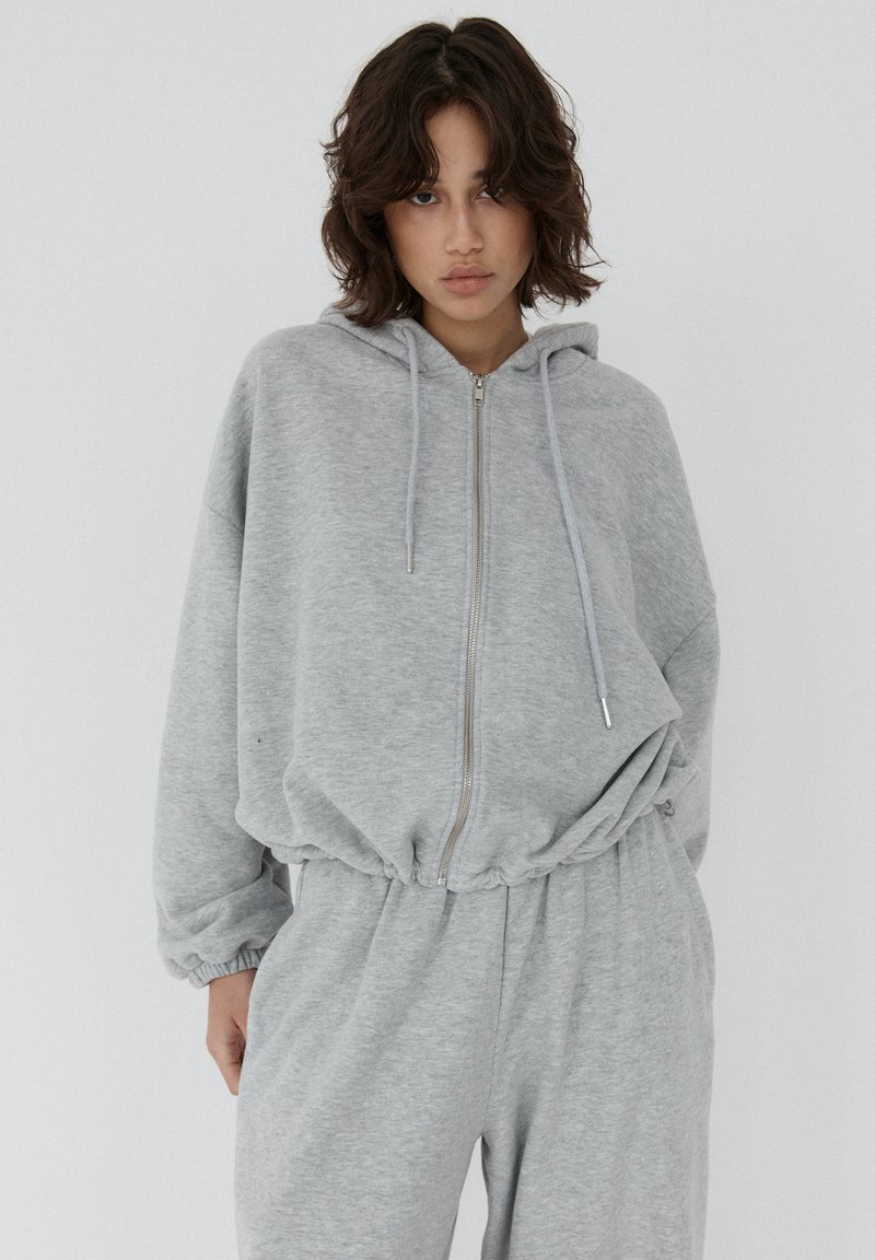 Młoda osoba z włosami średniej długości, ubrana w jasnoszary rozpinany hoodie i pasujące dresy, na jednolitym tle.