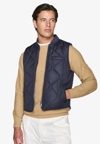 Marineblau gestepptes Puffer-Weste mit Reißverschluss, leichtem Design, hohem Kragen und Seitentaschen, über einem beigen Strickpullover getragen.