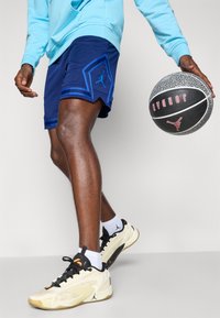 Pantalones cortos marinos con detalles azules y un diseño de diamante, combinados con zapatillas beige y calcetines blancos, sosteniendo un balón de baloncesto texturizado.