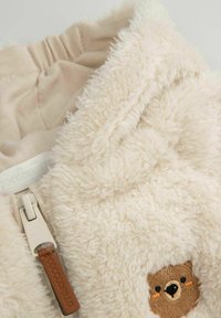 Kremowy polarowy hoodie z miękką, kudłatą fakturą, elastycznym pasem i haftem w kształcie twarzy misia z przodu.