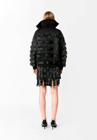 Chaqueta de plumas negra con detalles de cintas en capas y una falda negra a juego con flecos verticales. La modelo lleva zapatos de tacón negros.