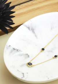 Collana in oro decorata con tre gemme nere su un piatto rotondo di marmo bianco con venature grigie, posto su uno sfondo chiaro.