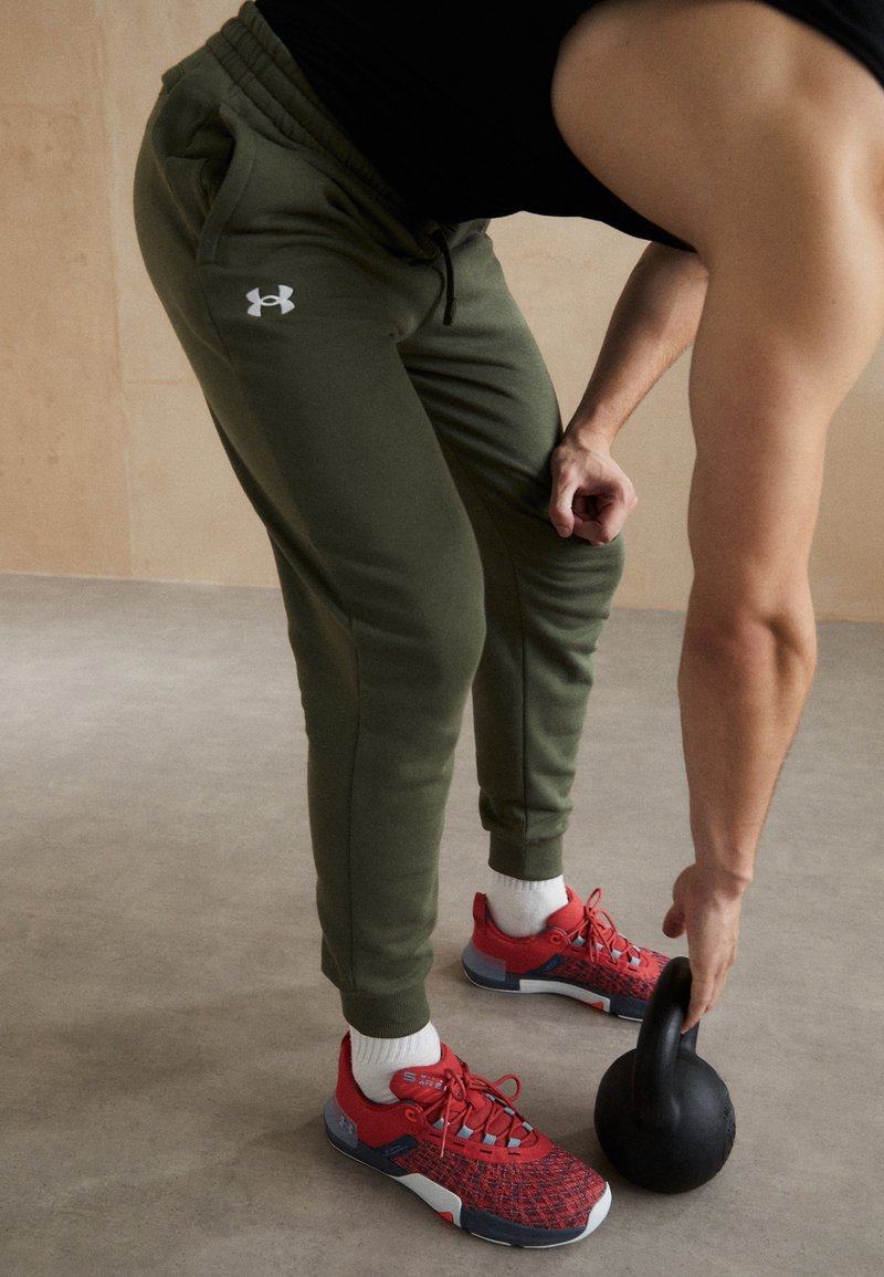 Grüne sportliche Jogginghosen mit elastischem Bund, weiße Logodetails. Rote Sportschuhe mit grauen Akzenten, während ein schwarzes Kettlebell gehoben wird.