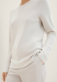 Maglia grigia chiara a maniche lunghe con sottili righe orizzontali e scollatura arrotondata, abbinata a pantaloni grigi chiari. Tessuto morbido, vestibilità rilassata.