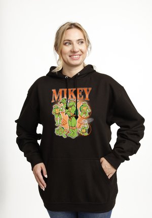 Kobieta uśmiechająca się, ubrana w czarną bluzę z kapturem z kilkoma zielonymi kreskówkowymi żółwiami i pomarańczowym napisem "MIKEY" z przodu.