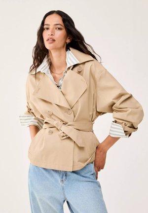 Donna con capelli lunghi e scuri che indossa un trench beige con cintura sopra una camicia a righe chiara e jeans azzurri, posando con una mano sul fianco.