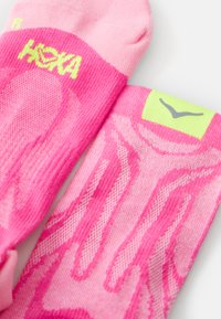 Pink athletic socks featuring a breathable mesh design, textured accents, and a light green label. Brand name "HOKA" displayed in bright yellow.