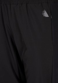 Crne sportske hlače napravljene od laganog materijala. Imaju elastični pojas, bočni patentni zatvarač i reflektirajući trokutasti logotip.