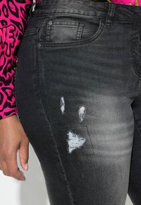 Jeans de mezclilla negra con un efecto desgastado, que presentan parches desgastados y un acabado texturizado. El primer plano muestra la colocación de la mano al lado de la costura lateral.
