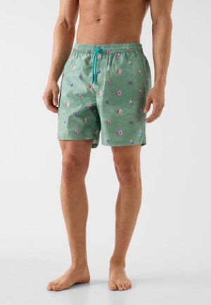 Männliches Model trägt grüne Badehose mit kleinen bunten Fruchtmustern, türkisfarbener Kordelzug, steht barfuß vor weißem Hintergrund.