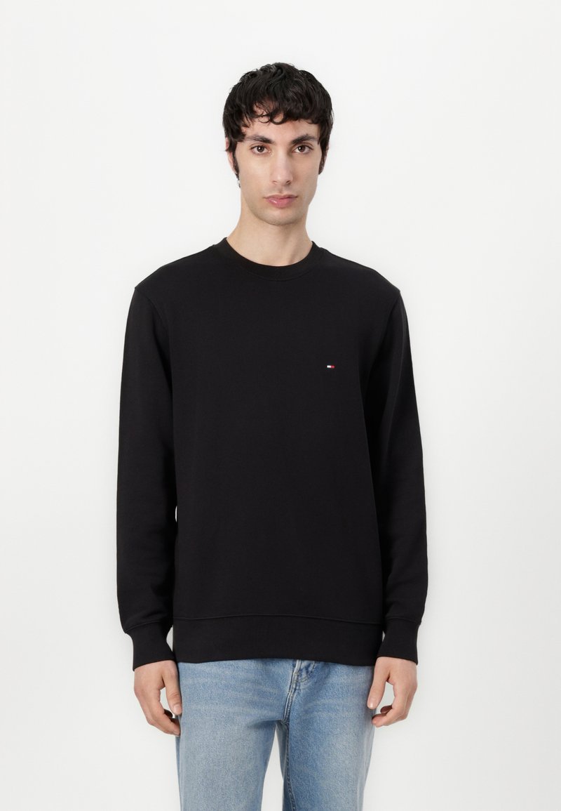Felpa nera con scollo rotondo, polsini e orlo a costine, caratterizzata da un piccolo logo multicolore sul lato sinistro del petto. Indossata con jeans azzurri chiari.
