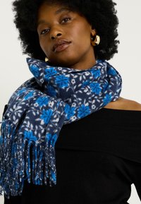 Mujer con afro oscuro que lleva pendientes de aro dorados, una blusa negra sin hombros y una bufanda azul con un patrón floral y flecos alrededor del cuello.