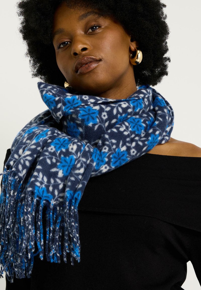 Mujer con afro oscuro que lleva pendientes de aro dorados, una blusa negra sin hombros y una bufanda azul con un patrón floral y flecos alrededor del cuello.