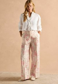 Biela ľanová blúzka s volánovým predným dielom, skombinovaná s krémovými nohavicami širokých nohavíc s ružovým kvetinovým vzorom. Neutrálne nazúvacie topánky.