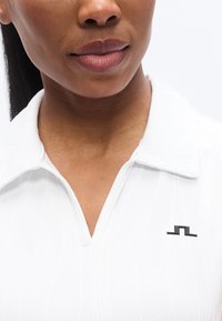 Λευκό πουκάμισο πόλο με κολάρο και υφή, με μαύρο λογότυπο στα αριστερά. Το κολάρο είναι δομημένο και το ύφασμα φαίνεται λείο.