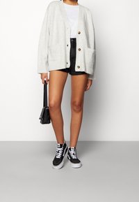 Cardigan de punto gris claro con dos bolsillos frontales, cierre de botones, usado sobre una camiseta blanca y pantalones cortos negros, combinado con zapatillas negras.