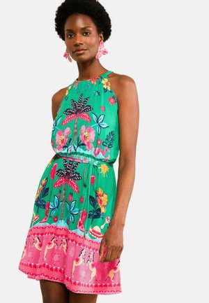 Nő élénk virág- és lómintás zöld ujjatlan ruhában, egyszerű világos háttér előtt áll.