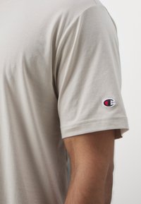 Beige bomull t-skjorte med korte ermer, med en liten brodert logo på ermet. Glatt tekstur og klassisk passform.