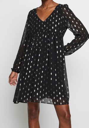 Czarna, przejrzysta sukienka z długim rękawem w srebrny diamentowy wzór, z dekoltem w kształcie litery V i luźnym, zwiewnym krojem, noszona przez osobę z jedną ręką na biodrze.