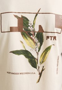 Camiseta de color beige claro con un gráfico de hojas en verde y blanco, con acentos marrones y texto que dice "NORTHWOODS WISCONSIN U.S.A." y "PETROL INDUSTRIES."
