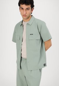 Short-sleeve, light green shirt with two front pockets and snap buttons, worn over a light beige shirt. Features a small logo patch.