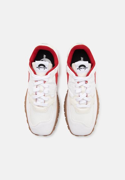 White sneakers with red accents, featuring a textured upper, lace-up closure, and a brown rubber sole. Notable brand logo on tongue.