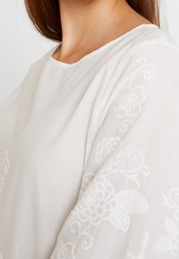 Blusa bianca con ricami floreali in un motivo texturizzato sulle maniche, con scollatura rotonda e vestibilità morbida.