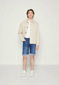 Casaco de linho bege, t-shirt branca, calções de ganga azuis com as bainhas dobradas, ténis brancos. Modelo posicionado ao centro contra um fundo simples.