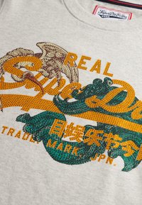 Grå T-shirt med korte ærmer, der har farverig dekorativ tekst med metaliske detaljer: orange, grønne og brune detaljer i forskellige skrifttyper og designs.