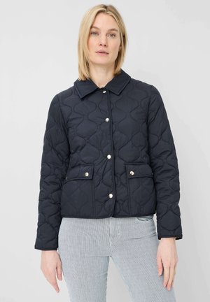 Woman with blonde hair wears a black quilted jacket with silver snap buttons and striped light pants against a plain white background.