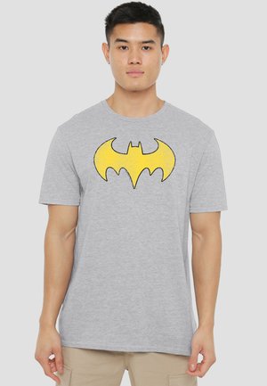 Young man wearing a light gray T-shirt with a large yellow Batman logo, standing against a plain light background.