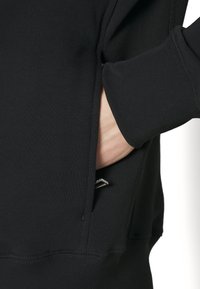 Main insérée dans la poche latérale d'un vêtement noir à manches longues avec un poignet côtelé et des détails de coutures visibles.