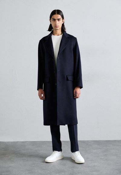 Lungo cappotto blu scuro realizzato in lana con colletto a scolla, due tasche frontali e chiusura con bottoni, abbinato a sneakers bianche e pantaloni.