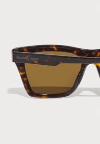 Ochelari de soare cu model broască țestoasă, cu lentile brunii, având un design robust și unghiular și un logo argintiu "MICHAEL KORS" pe sprijin.