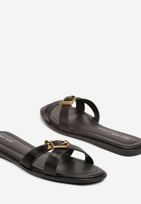 Sandales en cuir noir à enfiler avec une semelle plate, présentant des sangles croisées et une boucle dorée sur le devant. Texture lisse.