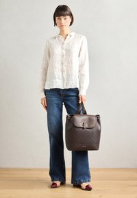 Un sac à dos en cuir marron au design structuré, avec un motif texturé et une fermeture à rabat. Porté avec une chemise en lin blanche et un jean évasé.