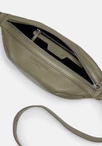 Sac en cuir vert olive avec une texture lisse, comportant un compartiment zippé et une étiquette intérieure de marque. Comprend une bandoulière fine amovible.