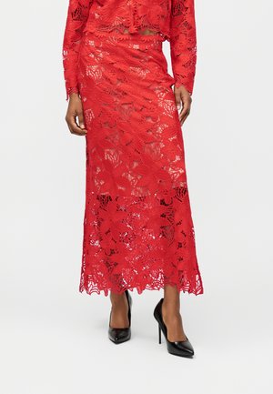 Jupe en dentelle florale rouge avec des détails transparents portée avec des talons hauts noirs à bout pointu, montrant le bas du torse et les jambes de la porteuse.