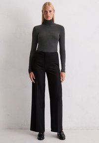 Frau steht vor einer weißen Wand, trägt einen dunkelgrauen Rollkragenpullover, schwarze weit geschnittene Hosen und schwarze Slipper, und blickt mit einem neutralen Gesichtsausdruck geradeaus.