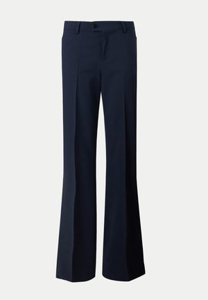 Femme debout portant un haut rose ajusté à manches courtes, un pantalon droit bleu marine et des chaussures à talons noirs, une main dans la poche.
